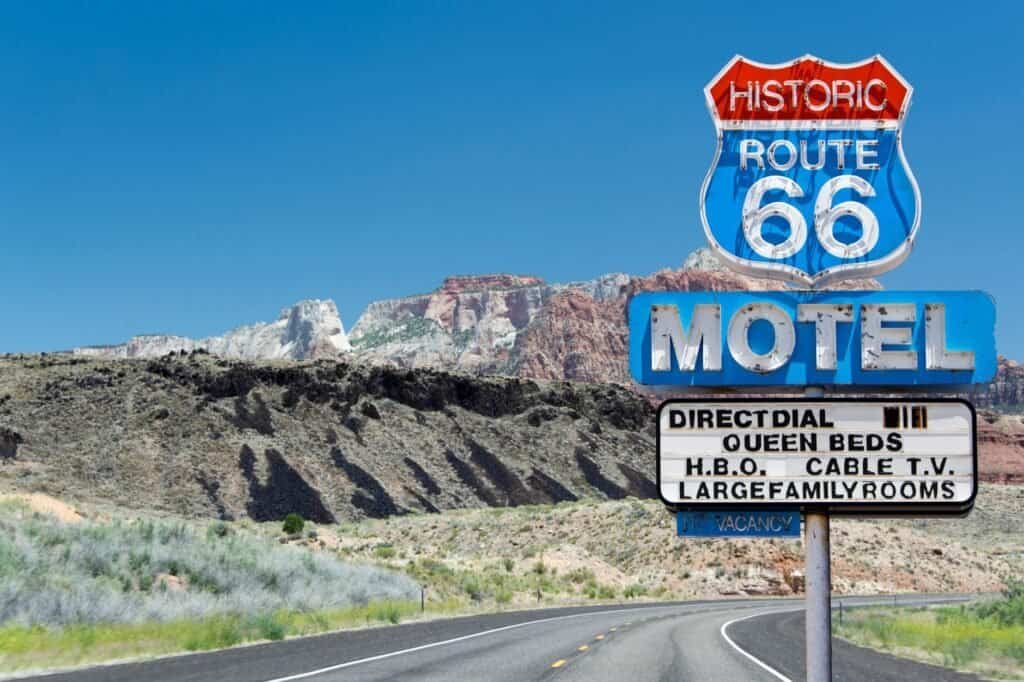 Route 66 motel sign by a winding road, nestled in a mountainous landscape under a clear blue sky—a classic backdrop for your summer adventure ideas.