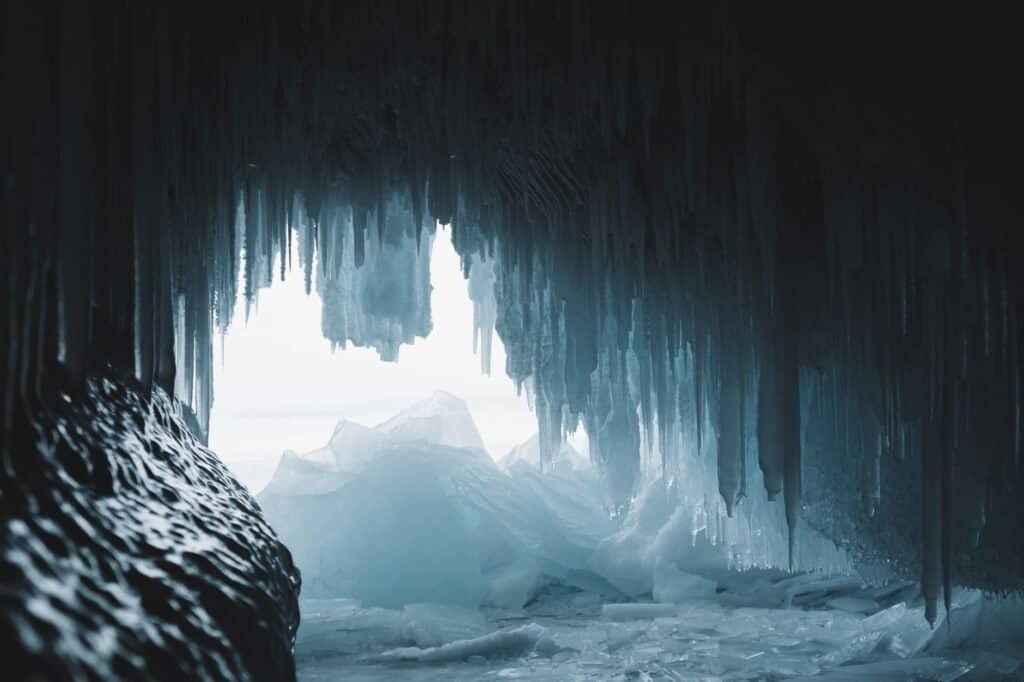 Explore the breathtaking beauty of an ice cave interior, where icicles hang from the ceiling, opening to a mesmerizing view of a snow-covered landscape and distant ice formations. This captivating scene offers inspiration for your summer adventure ideas in an unexpected winter wonderland.