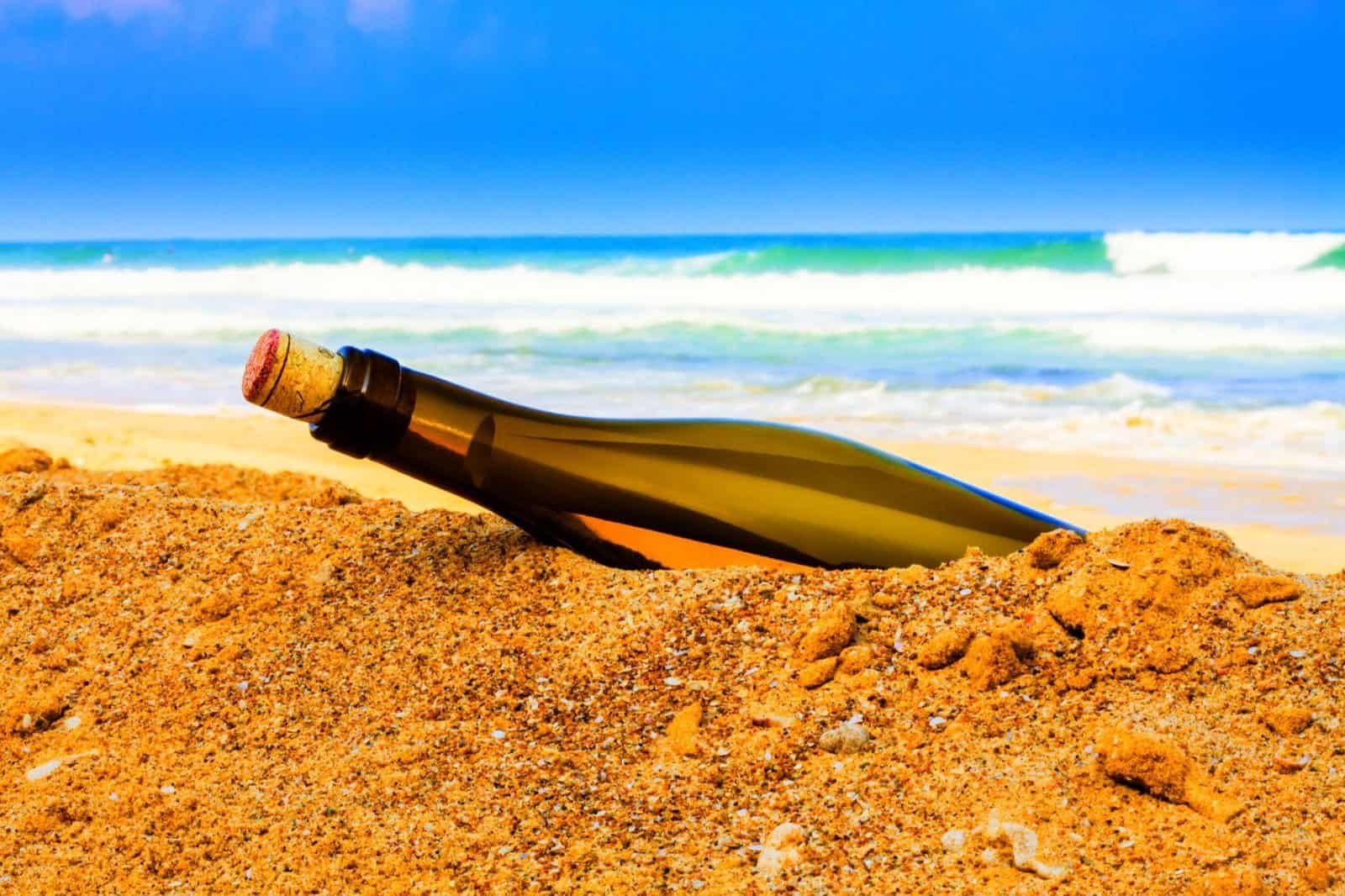 A corked glass bottle, perhaps a message in disguise, rests partially buried in the sand on a sunlit beach. With waves gently lapping and a blue sky stretching ahead, it invites thoughts of summer adventure ideas waiting to unfold.