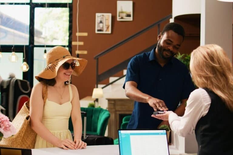 A woman in a sun hat and dress stands with a man at the hotel reception desk. As he hands a card to the receptionist, they seek cheap accommodation options. Behind them, a computer and elegant decor suggest smart choices for those traveling on a budget.