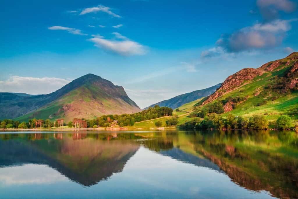 Discover spring adventure ideas as a serene lake reflects surrounding green mountains and trees under a clear blue sky with a few clouds.