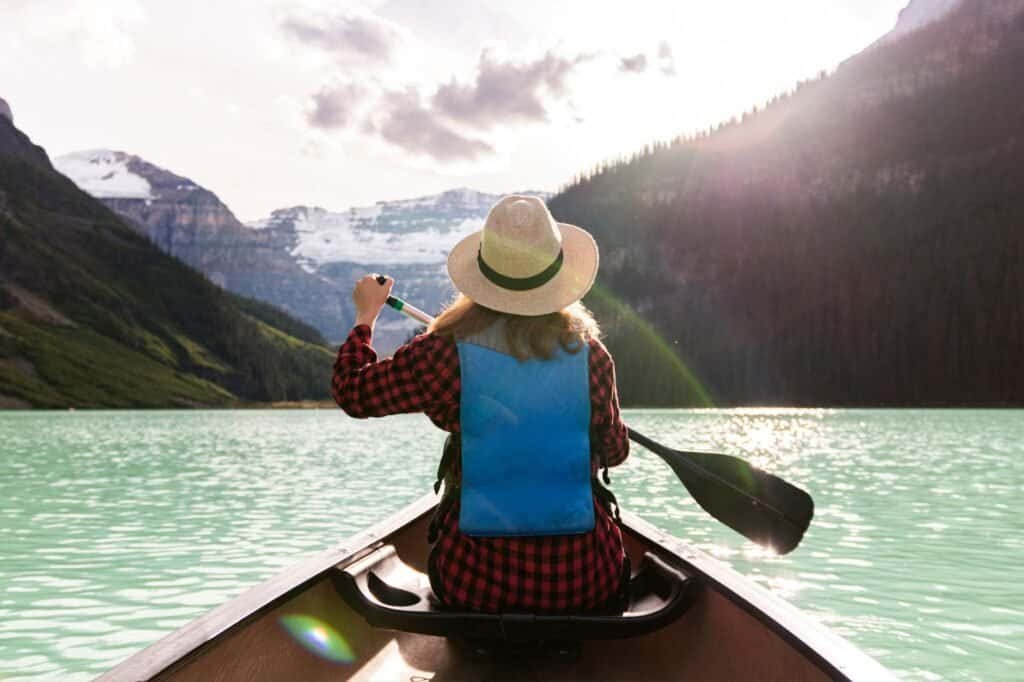 A person wearing a hat and life vest is paddling a canoe on a turquoise lake, surrounded by mountains and trees under a cloudy sky—an ideal spring adventure idea.