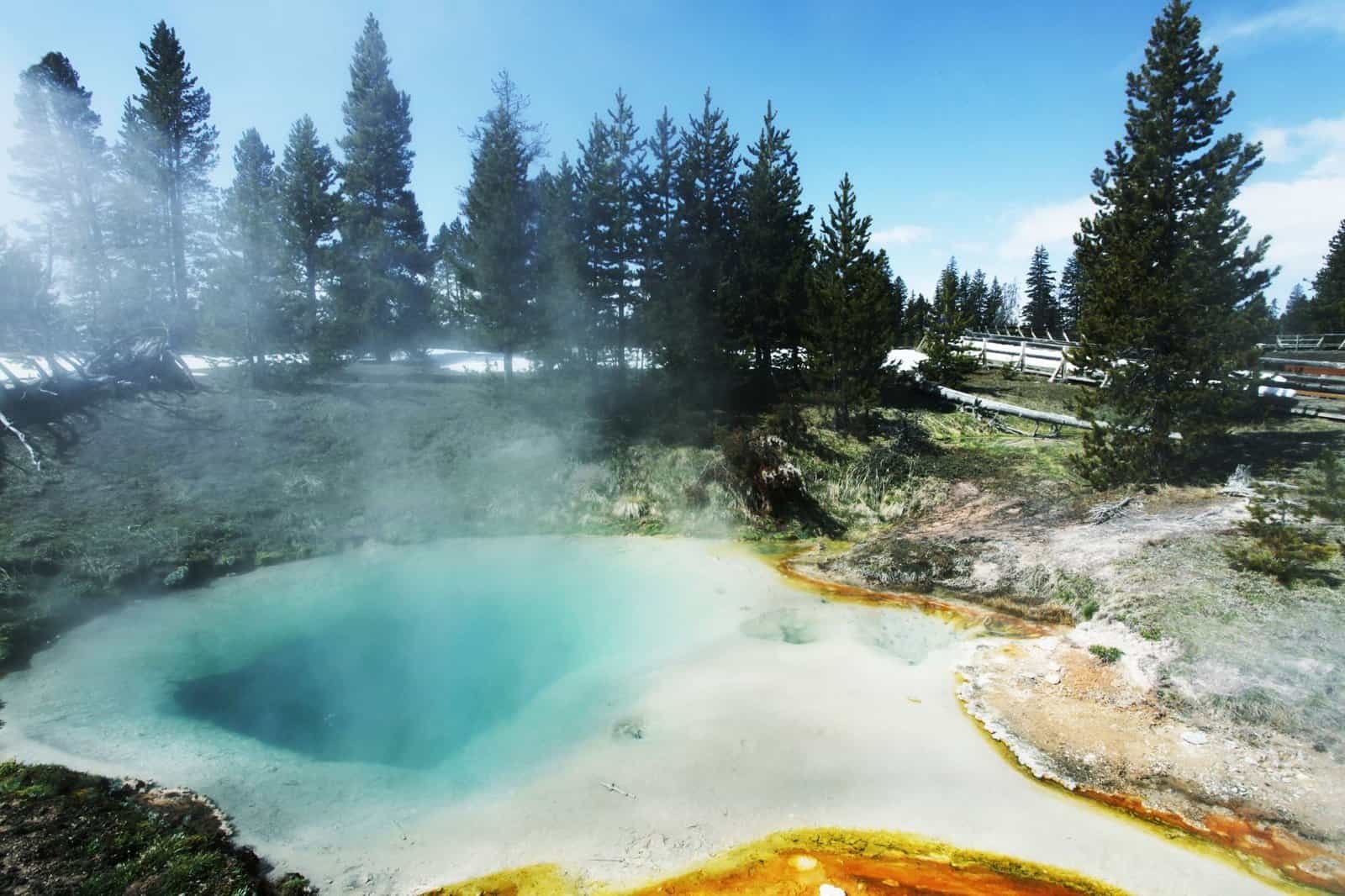 A hot spring adventure awaits with steaming blue water, nestled among trees and a wooden walkway under a clear sky.