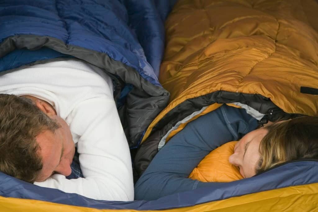 Two people are cozily sleeping side by side in budget sleeping bags, one blue and one yellow. They are facing each other.