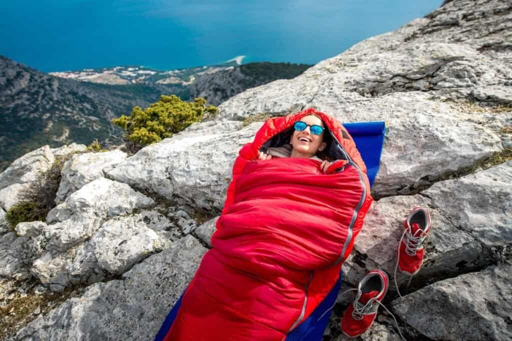 A person cozily nestled in a budget sleeping bag lies on rocky terrain, with red shoes placed nearby, enjoying a stunning view of the lake and distant town.