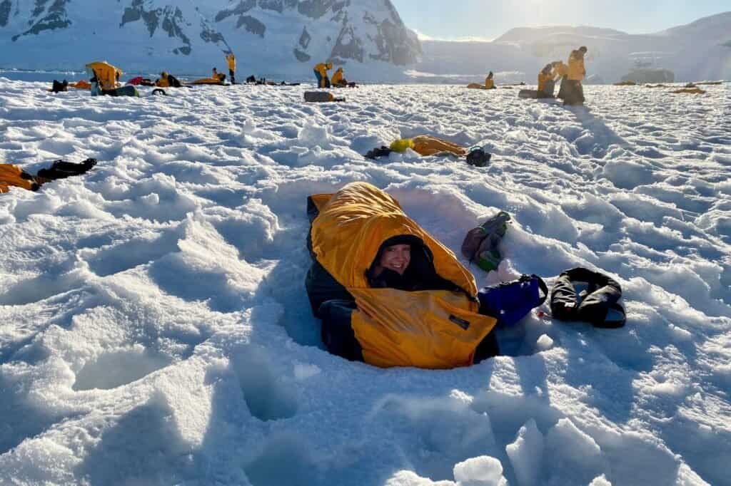 A person nestles in a cozy yellow budget sleeping bag on snowy terrain, encircled by snow-covered mountains and others in similar bags.