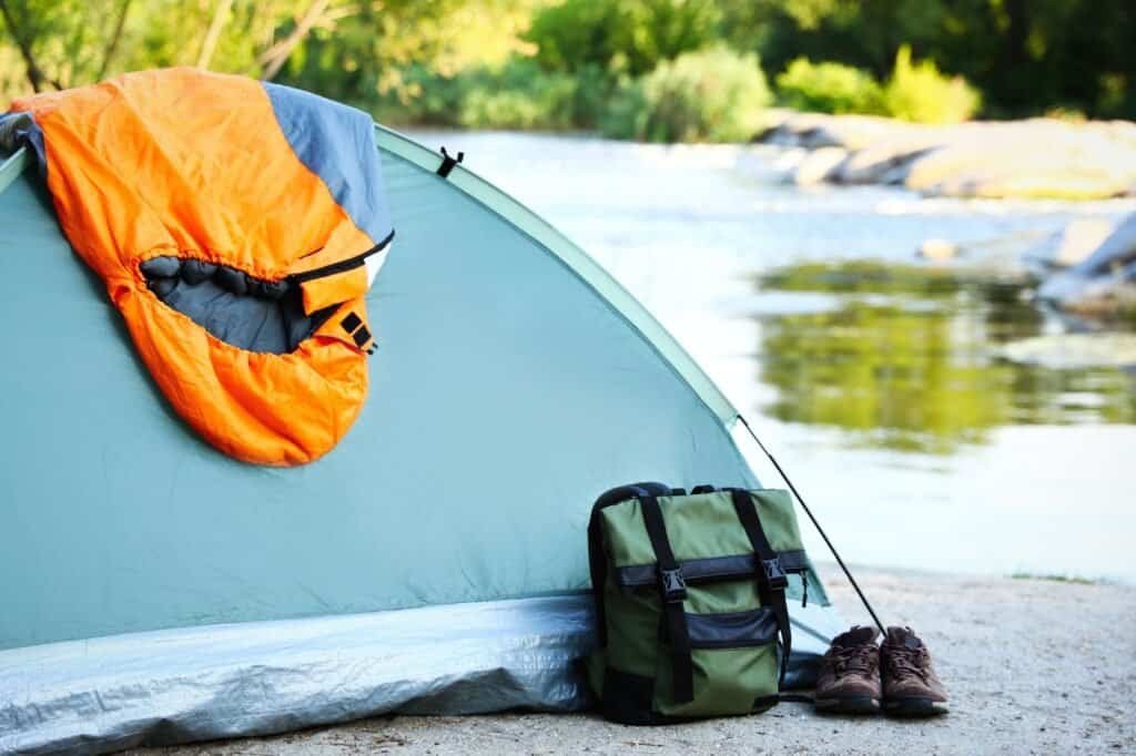 A green tent with a budget sleeping bag draped over it, a green backpack, and brown shoes is set up by a tranquil river, surrounded by rocks and greenery in the background.