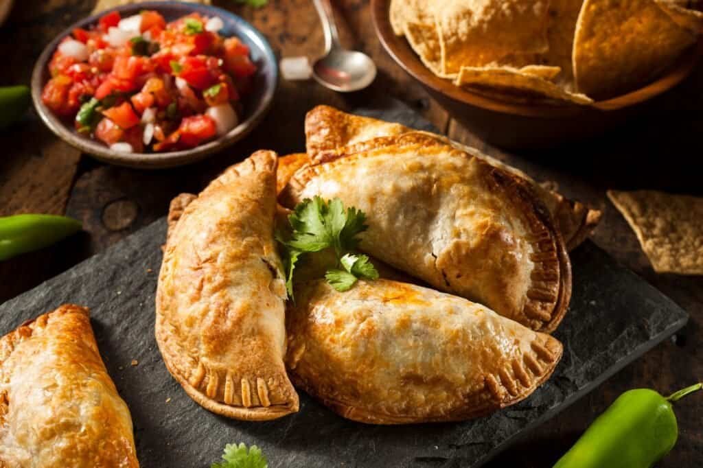 A slate platter with baked empanadas, garnished with cilantro, captures the essence of street food travel. In the background, a small bowl of salsa and a bowl of tortilla chips accompany the scene, while green peppers are artfully scattered around.