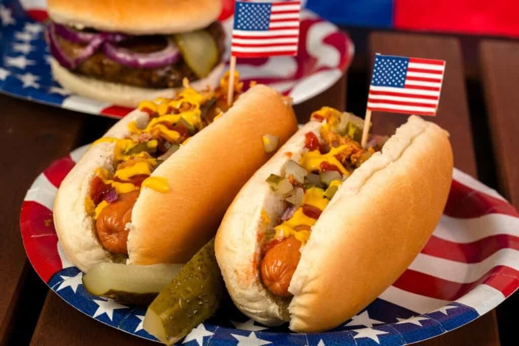 Two hot dogs in buns with mustard and relish are decorated with small American flags on a red, white, and blue plate—perfect street food for your travel adventures. In the background, a juicy hamburger awaits.