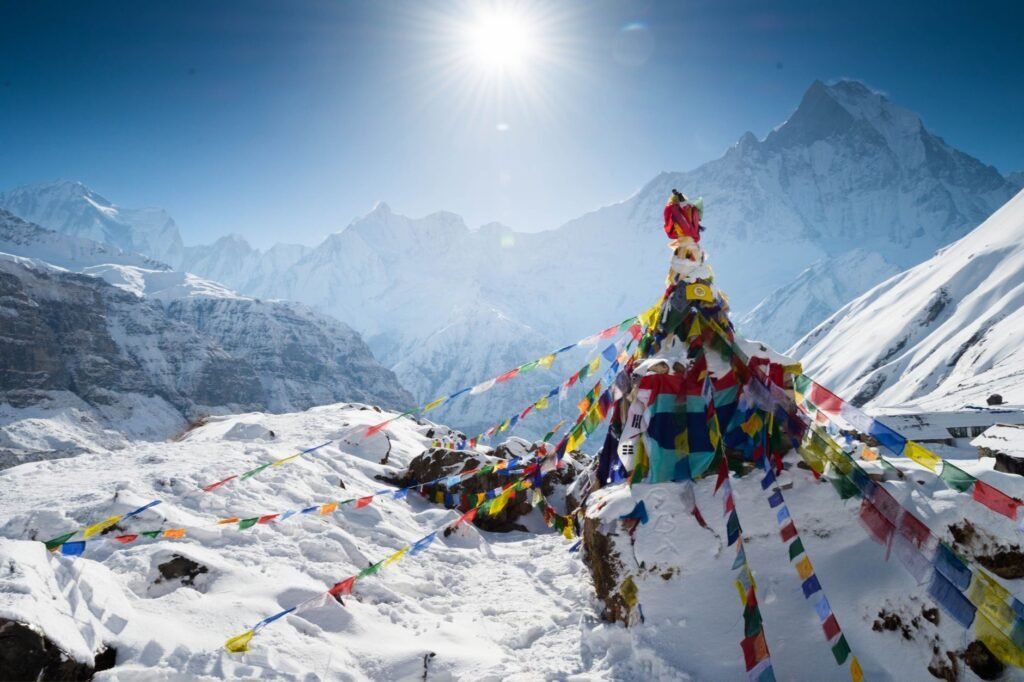 Vibrant prayer flags flutter on a snow-covered peak, with the bright sun casting a warm glow over distant mountains. Discover this enchanting scene among cheap places to travel, where beauty awaits at every turn.