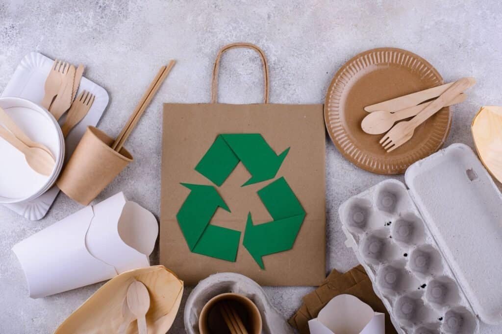 A recycling symbol on a paper bag surrounded by various biodegradable items like plates, cups, utensils, and egg cartons on a textured surface—ideal for those backpacking on a budget yet keen to stay eco-friendly.