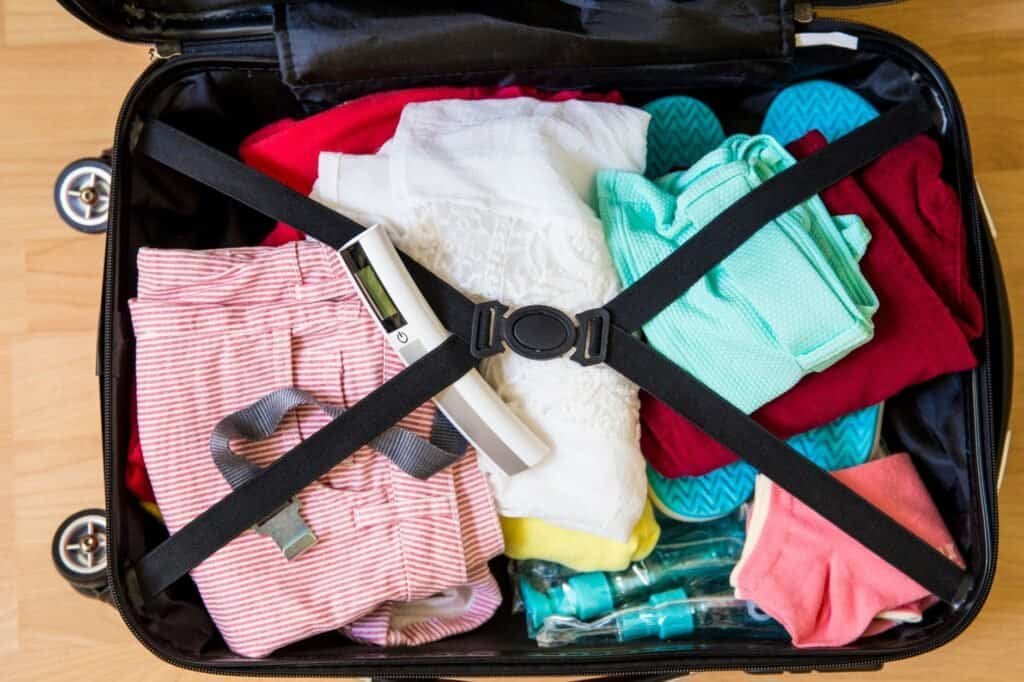 Open suitcase packed with neatly folded clothes, including shirts and swimwear, perfect for backpacking on a budget. A digital thermometer and travel bottles are secured with suitcase straps.