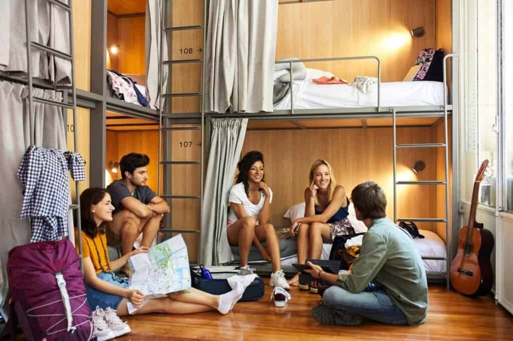A group of people sits and talks in a hostel dorm room, surrounded by bunk beds, luggage, and a guitar—the perfect setting for those backpacking on a budget.