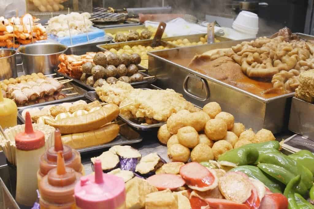 Assorted street food items, perfect for backpacking on a budget, include sausages, skewers, steamed buns, vegetables, and sauces all displayed invitingly on a vendor's stall.