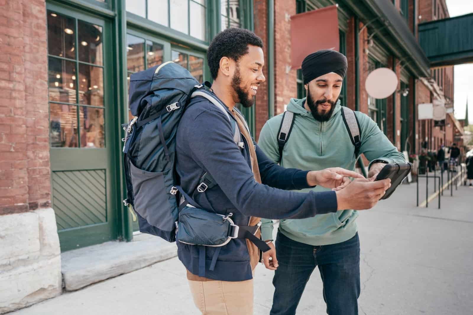 Two men with backpacks, possibly backpacking on a budget, examine a tablet while standing outside near brick buildings.