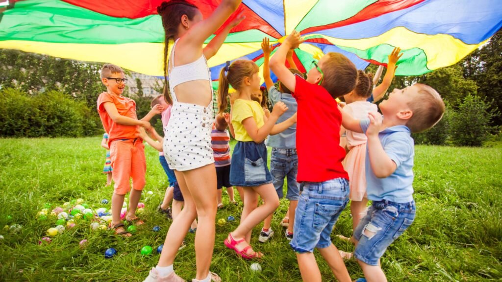 Children playing under a multicolored parachute on a grassy field, scattered plastic balls adding to the fun—a perfect scene of budget family travel tips come to life.