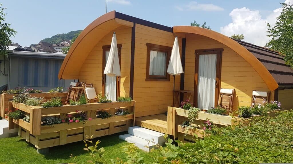 A small wooden cabin with a curved roof, surrounded by flower boxes and greenery; two deck chairs are visible near the entrance—perfect for families seeking budget-friendly retreats while traveling with kids.