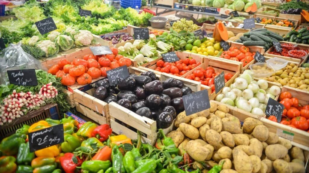 A colorful variety of fruits and vegetables displayed in wooden crates at a market, perfect for those seeking budget-friendly family meals. The produce includes tomatoes, eggplants, potatoes, peppers, onions, and more, with price tags visible. Consider this stop on your next budget-friendly trip!.