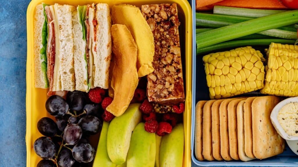 A yellow lunchbox packed with budget-friendly delights for a family adventure: sandwiches, melon slices, a granola bar, grapes, raspberries, apple slices, celery sticks, corn, crackers, and hummus. Perfect for fueling your journey while exploring travel tips on how to travel with kids on a budget!.