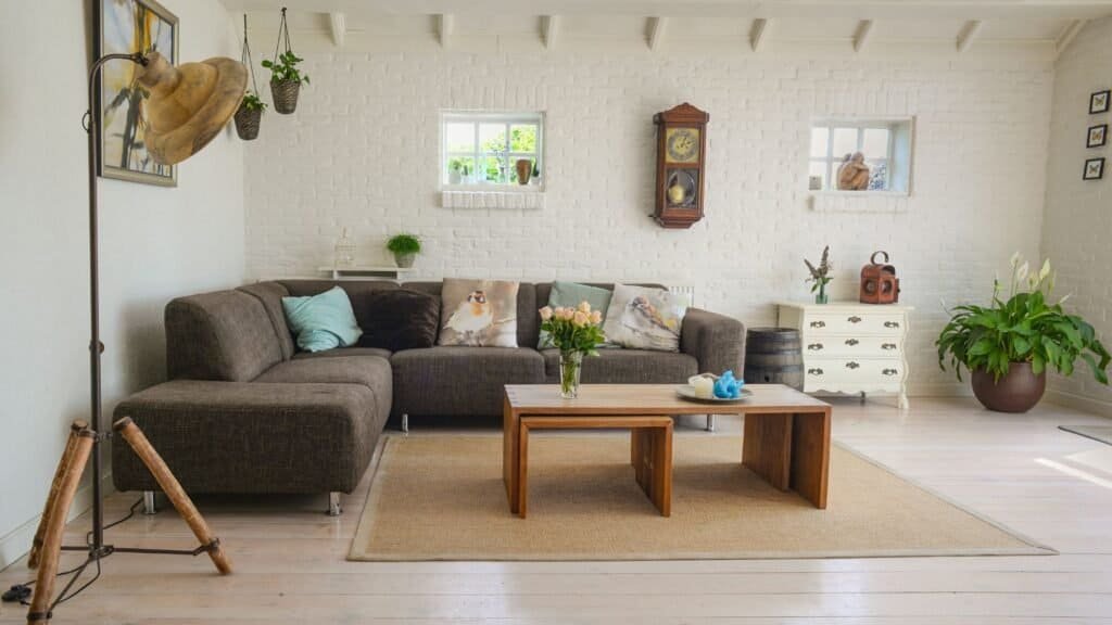 Cozy living room with a brown sectional sofa, wooden coffee table, and floral chairs—perfect for relaxing after a day of family travel. Decor features a wall clock, potted plants, and a floor lamp on light wood flooring.