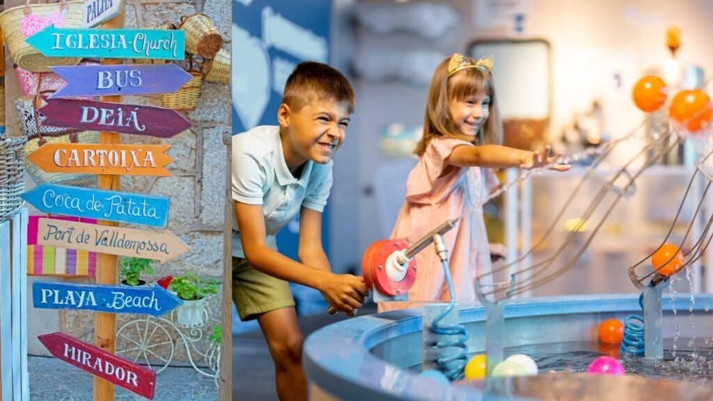 Two children play with water and colorful balls at an interactive exhibit. A colorful directional sign is on the left, pointing to spots like a church and beach—perfect for families exploring travel hacks that make every outing a breeze.