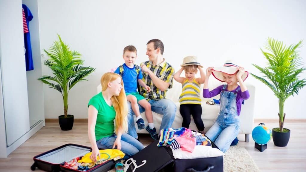 A family of five follows a packing guide for budget family travel in their bright living room with two potted plants. The children try on hats as the mom and dad arrange clothes, ensuring everything fits snugly into their suitcases.