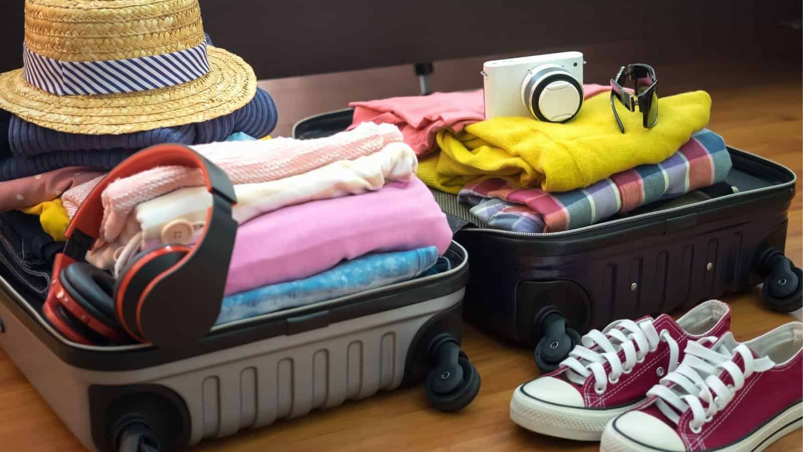 Two open suitcases on a wooden floor showcase a budget family travel packing guide, filled with colorful clothes, a straw hat, camera, headphones, watch, and vibrant red sneakers beside them.