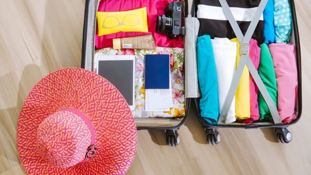 Open suitcase filled with neatly organized clothes, a camera, iPad, passport, and sunglasses—perfect essentials for someone planning how to visit Disneyland on a budget. A pink sunhat sits beside it on the wooden floor.