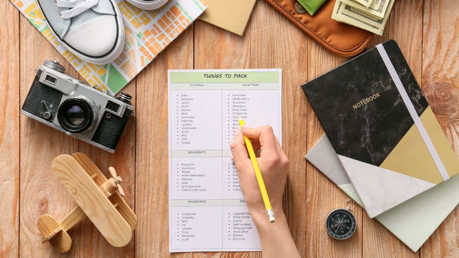 A person writes on a packing checklist surrounded by a camera, shoes, notebook, map, compass, cash, and a wooden toy plane on a wooden table.