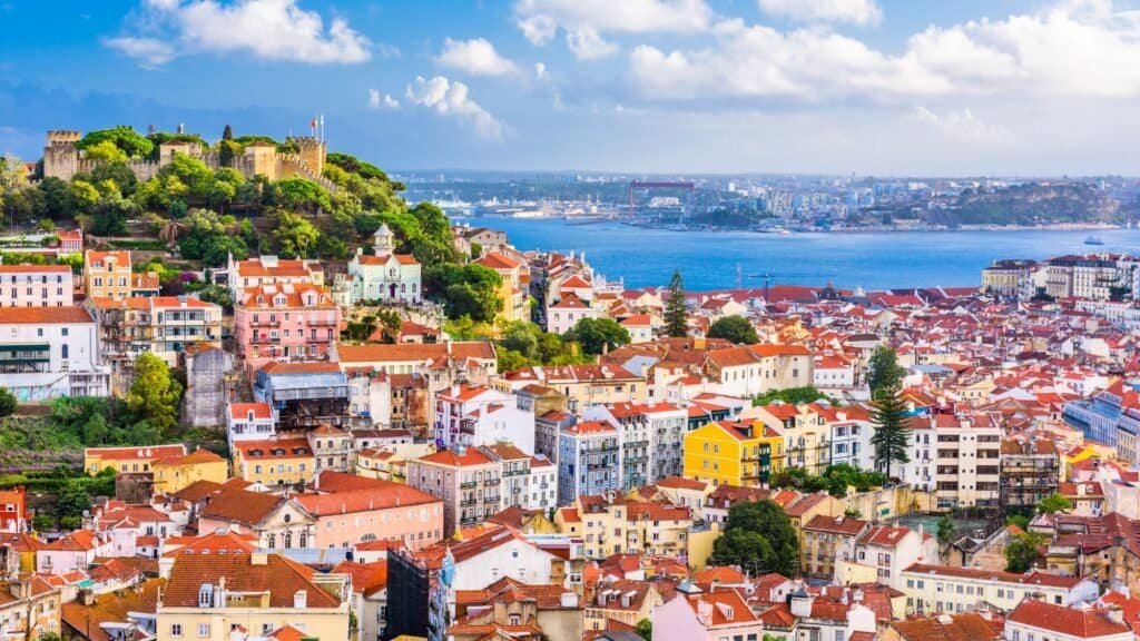 A panoramic view of Lisbon with colorful buildings, red-tiled roofs, a hilltop castle, and the Tagus River under a partly cloudy sky—perfect for solo female budget travel adventures.
