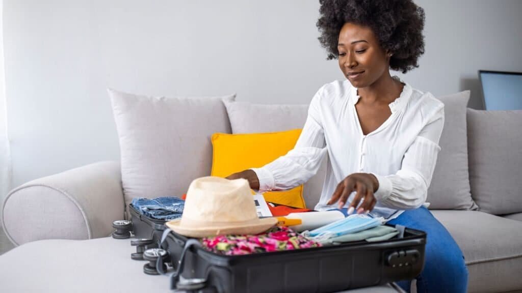 A woman sits on a couch, packing clothes and accessories into an open suitcase—getting ready for her next adventure in budget solo female travel.