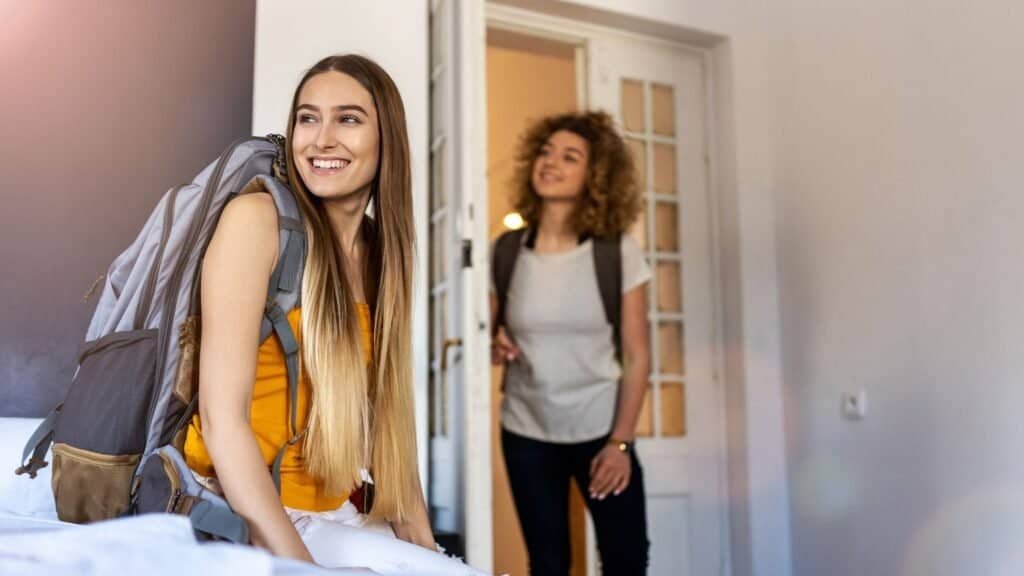 Two young women with backpacks are in a bright room; one sits smiling on the bed while her friend enters, capturing the joy and excitement of solo female travel adventures.