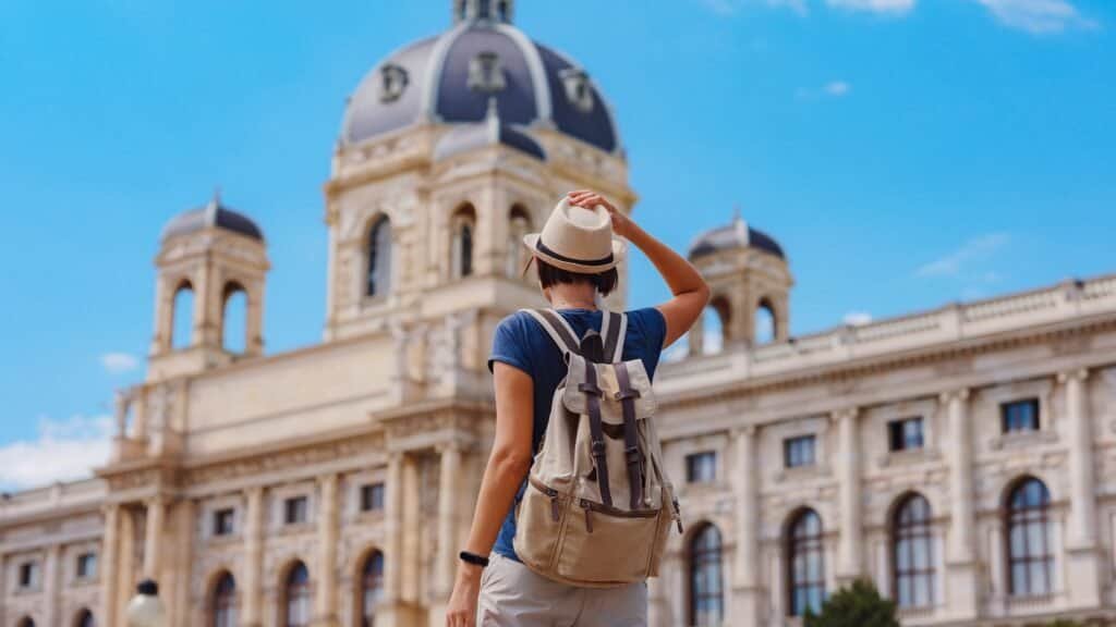 A solo female traveler wearing a hat and backpack stands in front of a large historic building with domes and towers under a clear blue sky, capturing the spirit of budget solo female travel.