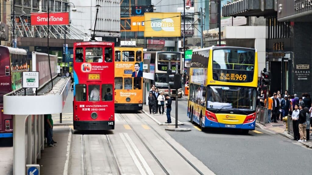 A busy street scene shows double-decker buses, trams, and pedestrians in a commercial area with shops and advertisements.